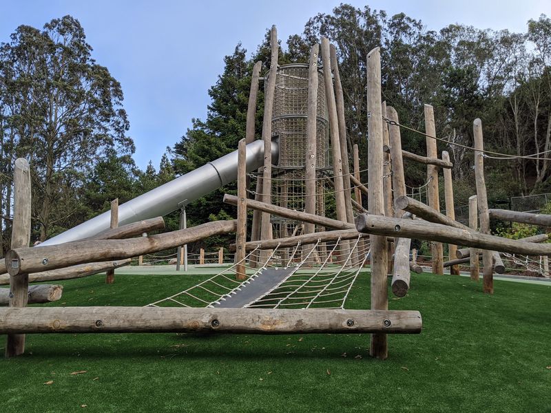 Climbing structure made of logs and rope net with a large tower and slide over green safety surface