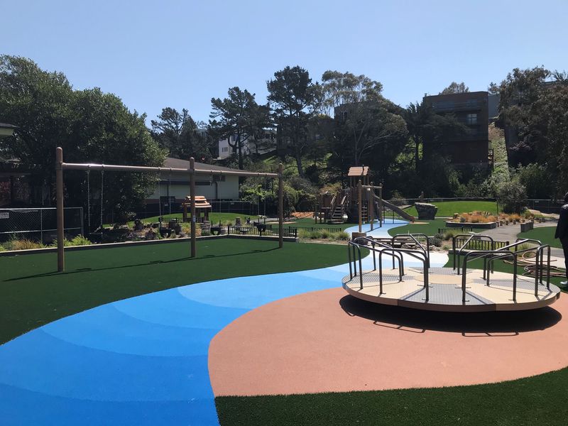 Merry go round and swing set on top of green surfacing with a blue strip through the middle.